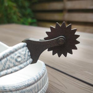 May include: A close-up shot of a metal spur featuring a star-shaped rowel. The spur is dark grey and connected to a white and blue patterned strap. The image is set against a wooden background, evoking a rustic or Western aesthetic.