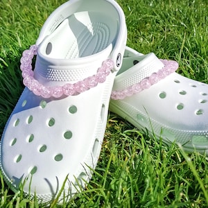 Op de afbeelding: Een paar witte klompen met een lichtroze kralenriem, liggend op groen gras. De klompen hebben meerdere ventilatiegaten en een textuurzool. De kralenriem voegt een vleugje kleur toe aan deze casual schoenen.