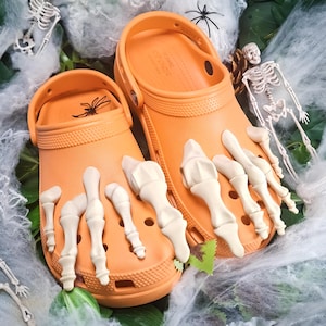 Könnte beinhalten: Ein Paar leuchtend orangefarbene Schlupfschuhe mit beigefarbenen, geformten Skeletthand-Anhängern aus Kunststoff, die auf der Oberseite befestigt sind. Die Schuhe haben einen strukturierten Fersenriemen und kreisförmige Belüftungslöcher.