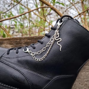 Könnte beinhalten: Ein schwarzer Schnürstiefel, der mit einer silberfarbenen Metallkette verziert ist. Das dekorative Kettenelement besteht aus zwei hängenden Gliedern und einem kleinen, strukturierten Schlangenanhänger, der mit einem Karabinerhaken befestigt ist.