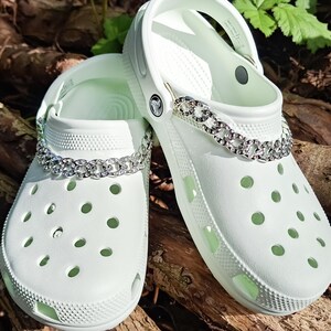 May include: A pair of white Crocs shoes with a silver chain embellishment. The shoes are made of a light green material and have multiple ventilation holes. The silver chain is adorned with clear rhinestones. The shoes are resting on a bed of brown wood.