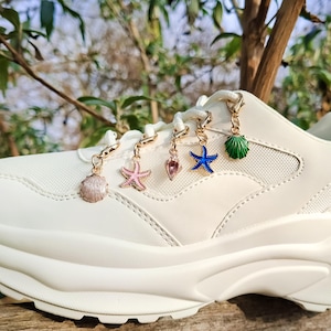 May include: Cream-coloured platform trainer decorated with seashell, starfish, and other sea-themed charms. The charms are in gold, pink, blue, and green. The shoe is set against a natural, outdoor background.