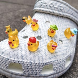Könnte beinhalten: Ein hellgrauer Crocs-Schuh, verziert mit mehreren gelben Enten-Anhängern. Die Anhänger tragen verschiedene Accessoires, darunter Hüte und Sonnenbrillen. Der Schuh steht auf einer grauen Oberfläche.