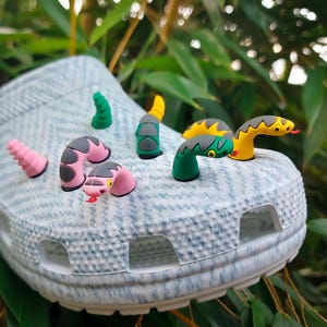 May include: A light grey and white marbled clog shoe decorated with several colourful, three-dimensional snake-shaped charms. The charms include various green, pink, yellow, and black snake heads and tails protruding from the shoe's ventilation holes.