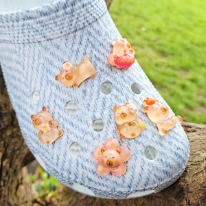 Könnte beinhalten: Ein hellblauer Clog-Schuh mit Denim-Textur, verziert mit mehreren kleinen, durchscheinenden Bären-förmigen Charms. Die Charms sind in verschiedenen Posen und weisen Brauntöne, Rosa und Orange auf. Der Schuh liegt auf einer braunen, strukturierten Oberfläche mit grünem Hintergrund.
