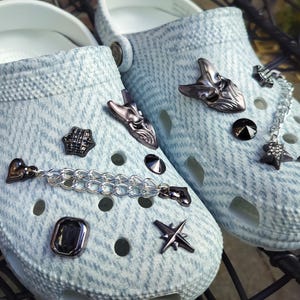 Könnte beinhalten: Hellblaue Clogs mit strukturiertem Muster, verziert mit dunklen Metallanhängern. Zu den Anhängern gehören eine Maske, Sterne, ein Herz und eine Kette. Die Clogs haben weiße Sohlen.