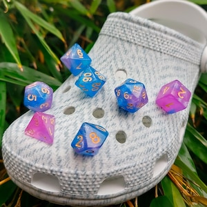 Peut inclure: Un sabot gris clair avec plusieurs dés colorés. Les dés sont bleus et violets avec des chiffres dorés. La chaussure est entourée de feuilles de bambou vertes.