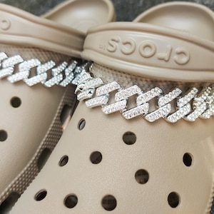 May include: Close-up of two beige Crocs sandals adorned with silver chain shoe charms. The charms are encrusted with small, sparkling rhinestones, adding a touch of glamour to the casual footwear. The Crocs have multiple holes.