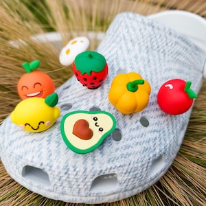 Puede incluir: Un par de zuecos blancos con una variedad de abalorios de frutas y verduras de colores. Los abalorios incluyen una fresa, una naranja, un limón, un aguacate, una manzana roja y un pimiento amarillo.