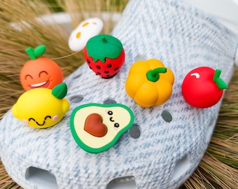 May include: A light gray textured clog shoe decorated with several colorful fruit and vegetable charms, including a smiling orange, a lemon, a strawberry, a yellow bell pepper, a red apple, and a halved avocado with a brown pit.