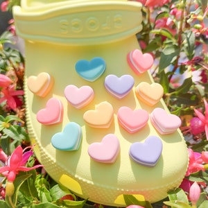 Puede incluir: Zapatilla amarilla Crocs con varios abalorios en forma de corazón de color pastel. Los abalorios son en tonos de rosa, azul, amarillo y morado.