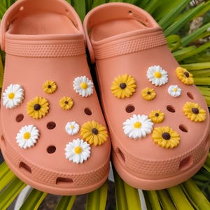 May include: A pair of peach-colored slip-on clogs featuring a series of circular ventilation holes on the upper surface. The clogs are decorated with multiple small, three-dimensional flower charms in shades of white and yellow, scattered across the front.