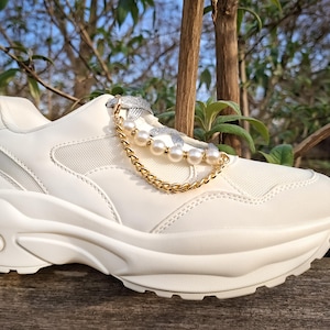 Könnte beinhalten: Weiße Plateau-Sneakers mit Obermaterial aus Mesh und Kunstleder. Die Sneakers haben eine goldene Kette und Perlenverzierung an den Schnürsenkeln. Die klobige Sohle sorgt für einen modernen Look. Vor einem natürlichen Hintergrund.