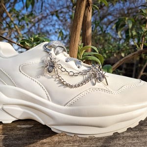 Op de afbeelding: Witte sneaker met dikke zool en mesh en leren panelen. De schoen heeft zilveren spinnenbedels die aan de veters hangen. De zool is dik en gestructureerd en het algehele ontwerp is modern en stijlvol.