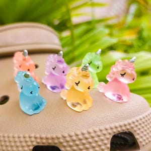 May include: A set of six small, translucent, colorful unicorn charms with silver-toned horns, each featuring a smooth, glossy finish and closed eyes, resting on the surface of a tan-colored, perforated foam clog shoe.
