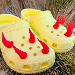 Könnte beinhalten: Ein Paar hellgelbe Clogs mit roten Teufelshorn-Anhängern. Die Clogs haben mehrere Löcher und eine strukturierte Oberfläche. Die roten Hörner sind oben an den Clogs befestigt.