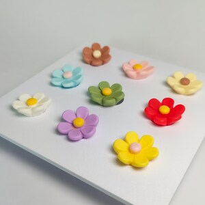 May include: A collection of nine small, multi-colored flower-shaped magnets arranged on a white square display board. Each flower features six rounded petals and a contrasting circular center in colors including red, yellow, blue, green, and pink.