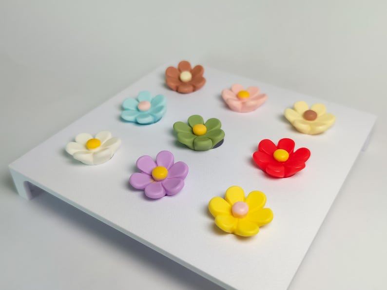 May include: A collection of nine small, multi-colored flower-shaped magnets arranged on a white square display board. Each flower features six rounded petals and a contrasting circular center in colors including red, yellow, blue, green, and pink.