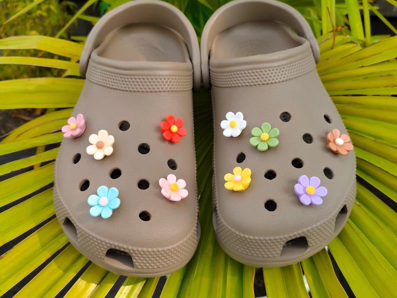 A pair of taupe clogs decorated with 10 colorful daisy flower shoe charms in assorted colors, displayed on tropical green leaves.