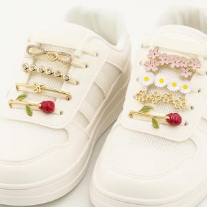 White sneaker decorated with various gold, crystal, and floral shoelace clips arranged horizontally across the laces.