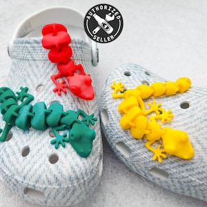 Könnte beinhalten: Zwei Paar bunte Gecko-Schuhcharms. Ein Paar ist grün und das andere gelb. Die Charms sind an einem Paar weißen Clogs befestigt.