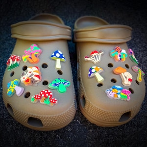 May include: A pair of tan slip-on clogs featuring a variety of colorful, small mushroom-shaped charms attached to the ventilation holes. The charms display diverse patterns and vibrant colors including red, blue, green, orange, and yellow.