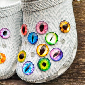 Könnte beinhalten: Ein Paar hellgraue, texturierte Clogs, die mit mehreren runden Cabochons verziert sind, welche realistische Tieraugenmotive in verschiedenen Farben wie Blau, Grün, Gelb, Orange, Rot und Violett mit vertikalen schwarzen Pupillen zeigen.