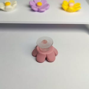 May include: A pink flower-shaped phone grip with a clear circular adhesive base. In the background, a white display stand holds three additional flower-shaped grips in white, light purple, and yellow, each with a central yellow dot.