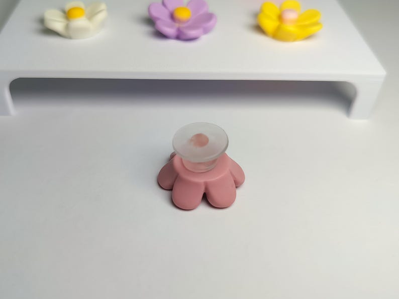 May include: A pink flower-shaped phone grip with a clear circular adhesive base. In the background, a white display stand holds three additional flower-shaped grips in white, light purple, and yellow, each with a central yellow dot.