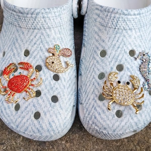 May include: A pair of light blue clogs with a herringbone pattern, adorned with decorative charms. Charms include a red crab, a bejeweled whale, a gold crab, and a seahorse. The clogs have multiple ventilation holes.