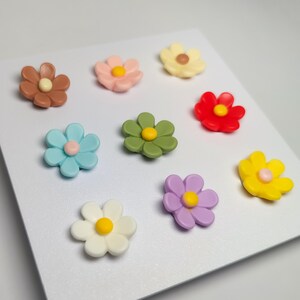 May include: A collection of nine small, multi-colored flower-shaped ornaments arranged in a grid. The flowers feature petals in shades of brown, pink, cream, red, light blue, olive green, yellow, white, and purple, each with a center dot.