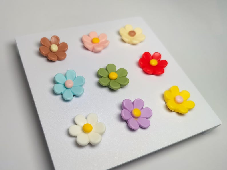 May include: A collection of nine small, multi-colored flower-shaped ornaments arranged in a grid. The flowers feature petals in shades of brown, pink, cream, red, light blue, olive green, yellow, white, and purple, each with a center dot.