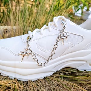 Könnte beinhalten: Ein weißer Plateau-Sneaker mit einer silbernen Kette und Stern-Anhängern. Die klobige Sohle und die Mesh-Details verleihen dem Schuh Struktur. Die Stern-Anhänger hängen an den Schnürsenkeln.