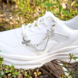 Op de afbeelding: Een witte sneaker met een zilverkleurige metalen ketting bevestigd aan de veters. Het decoratieve accessoire bevat twee kleine vlinderbedels met gedetailleerde vleugels, verbonden door een fijne schakelketting die over de schoen heen valt.