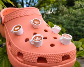 May include: A peach-colored foam clog shoe decorated with five miniature white coffee cup charms. Each charm features a brown liquid interior with detailed white latte art designs, including hearts and rosettes, securely attached to the shoe's surface.