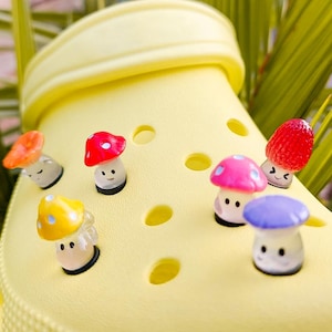 May include: A bright yellow foam clog shoe decorated with six small, colorful mushroom-shaped charms. The charms feature various cap colors including orange, red, yellow, pink, and purple, each with a small, simple face printed on the stem.