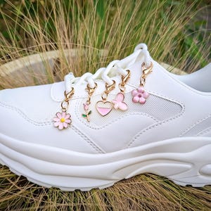 Könnte beinhalten: Ein weißer Sneaker mit dicker Sohle, verziert mit fünf goldfarbenen Charms. Die Charms zeigen eine rosa Gänseblümchen, eine rosa Rose, ein rosa Herz, einen rosa Schmetterling und eine rosa Blume. Der Schuh steht vor einem Hintergrund aus hohem Gras.