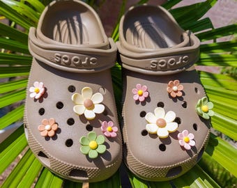 May include: A pair of taupe-colored slip-on clogs featuring a perforated design and a pivoting heel strap with the brand name Crocs embossed on the side. The shoes are decorated with various small, colorful plastic flower charms in shades of pink, yellow, white, peach, and light green.