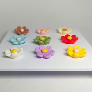 May include: A set of nine small, multi-colored flower-shaped magnets arranged in a three-by-three grid on a white rectangular display board. The flowers feature various colors including brown, pink, cream, blue, green, red, white, purple, and yellow.