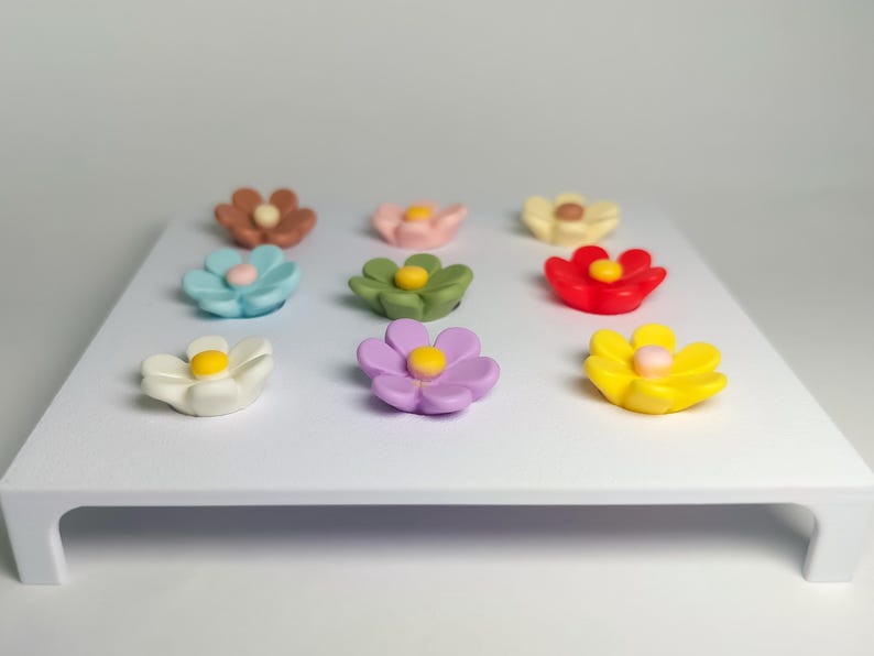 May include: A set of nine small, multi-colored flower-shaped magnets arranged in a three-by-three grid on a white rectangular display board. The flowers feature various colors including brown, pink, cream, blue, green, red, white, purple, and yellow.
