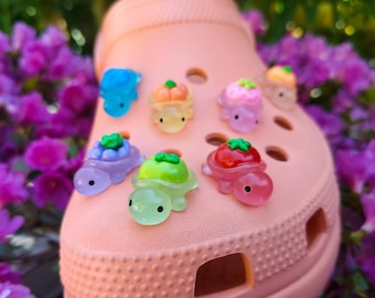 Puede incluir: Un zueco de color melocotón decorado con varios pequeños dijes translúcidos en forma de tortuga. Cada tortuga presenta una fruta o verdura de diferente color en su caparazón, incluyendo una baya azul, una calabaza naranja, una flor rosa, una lima verde y un tomate rojo.