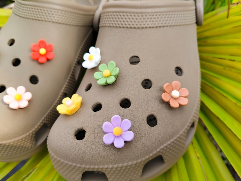 A pair of taupe clogs decorated with 10 colorful daisy flower shoe charms in assorted colors, displayed on tropical green leaves.
