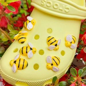 Puede incluir: Zuecos amarillos con abalorios de abejas. Los zuecos tienen una superficie texturizada y están decorados con varios abalorios de abejas en diferentes posiciones.