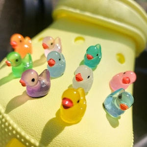 May include: A bright yellow clog shoe decorated with ten small, translucent, multi-colored duck-shaped charms. The ducks are arranged across the surface of the shoe and feature vibrant colors including orange, green, purple, blue, pink, and yellow.
