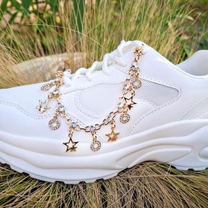 Puede incluir: Una zapatilla blanca con plataforma y una cadena dorada con colgantes de estrellas y círculos. La cadena está colocada sobre los cordones y el lateral del zapato. La zapatilla está sobre un fondo de hierba alta.