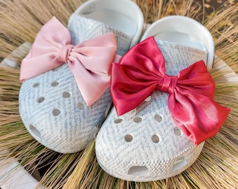 May include: A pair of light gray slip-on clogs with a textured herringbone pattern. The left clog features a large, smooth, pale pink satin bow, while the right clog features a large, smooth, deep red satin bow attached to the top surface.