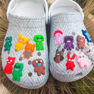 May include: A pair of light gray clogs decorated with colorful cartoon character charms. The charms are various colors and shapes, including green, yellow, orange, red, purple, brown, and pink. The clogs have white straps and are set against a background of green and brown grass.