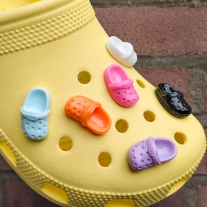 Puede incluir: Un zueco amarillo adornado con coloridos dijes. Los dijes incluyen diseños de zapatos en miniatura en azul, naranja, rosa, negro, blanco y morado. El zapato está sobre un fondo de ladrillos.