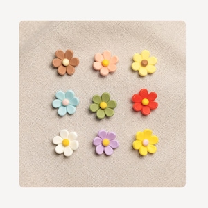 May include: A set of nine small flower-shaped embellishments arranged in a grid. Each piece features five rounded petals and a contrasting center dot. The colors include brown, peach, yellow, light blue, olive green, red, white, purple, and bright yellow.