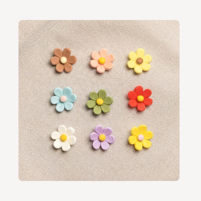 May include: A set of nine small flower-shaped embellishments arranged in a grid. Each piece features five rounded petals and a contrasting center dot. The colors include brown, peach, yellow, light blue, olive green, red, white, purple, and bright yellow.